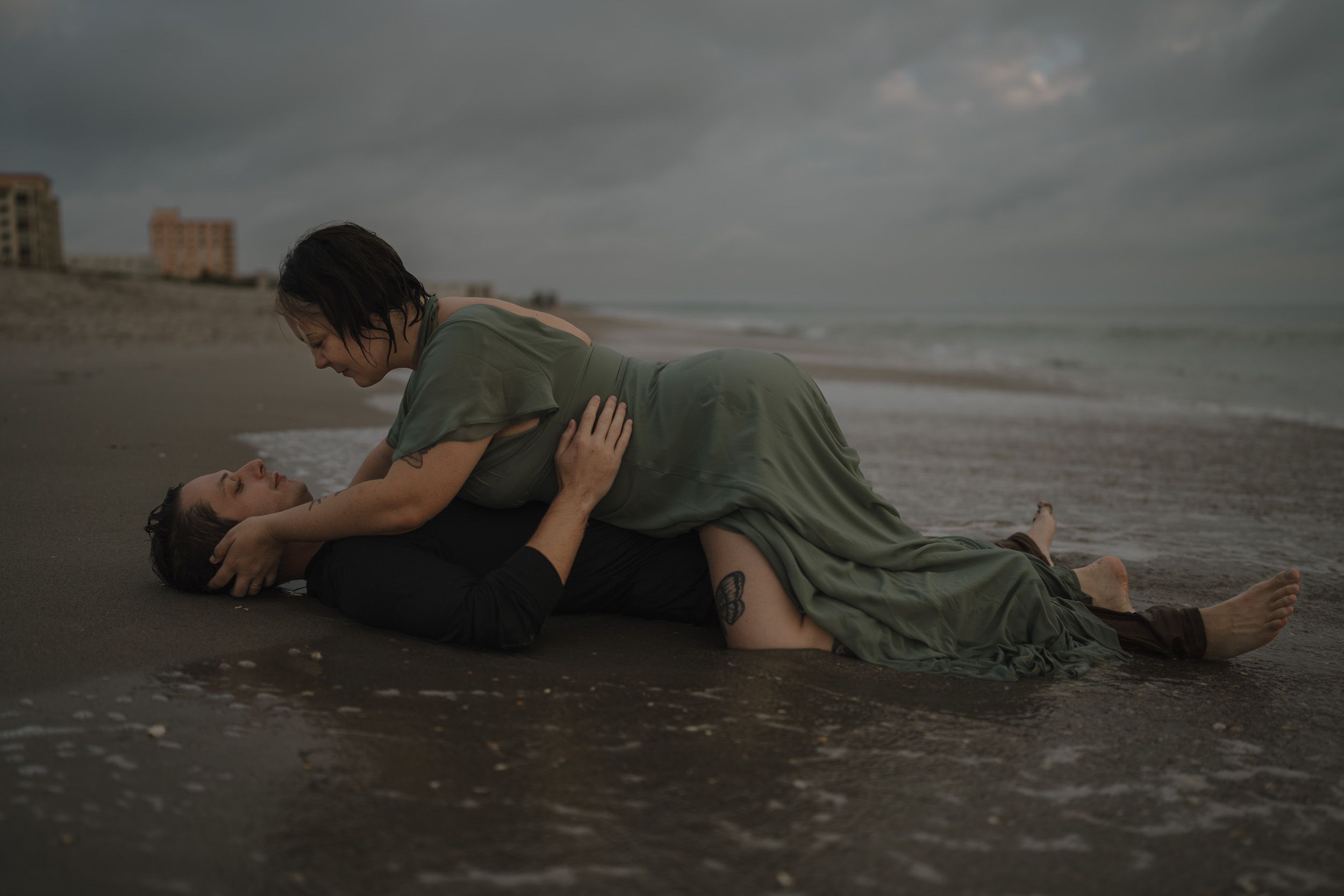 A couple lay near ocean waves as water washes over them at sunrise creating intimate photos. moody couples session in Cocoa Beach, Florida