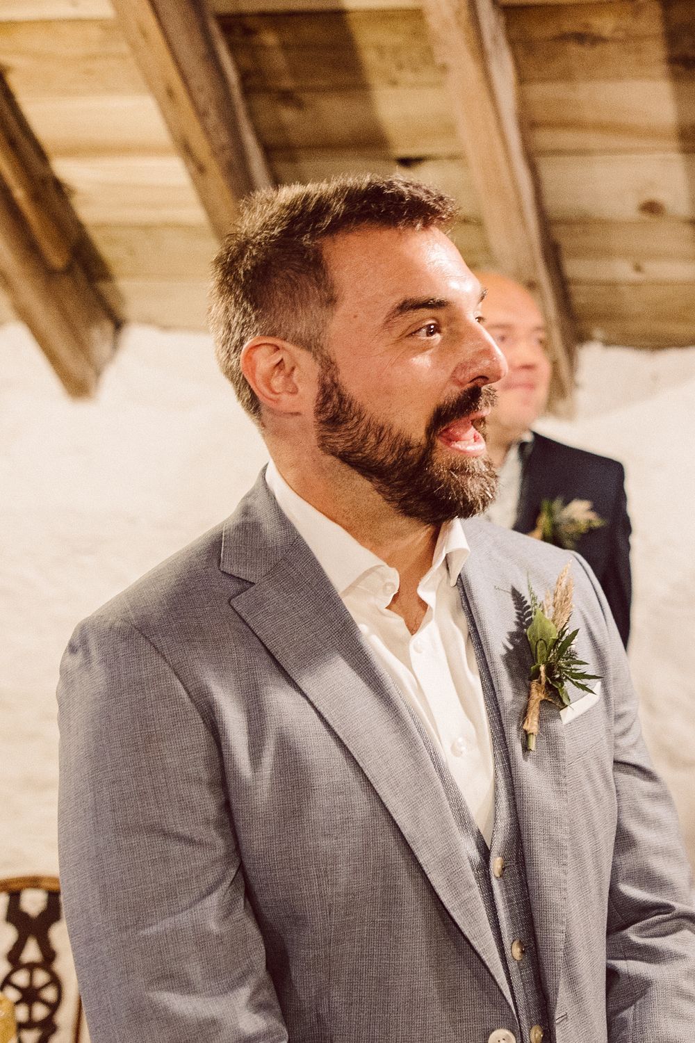 A groom in a gray suit and white shirt smiles during a rustic barn wedding ceremony.