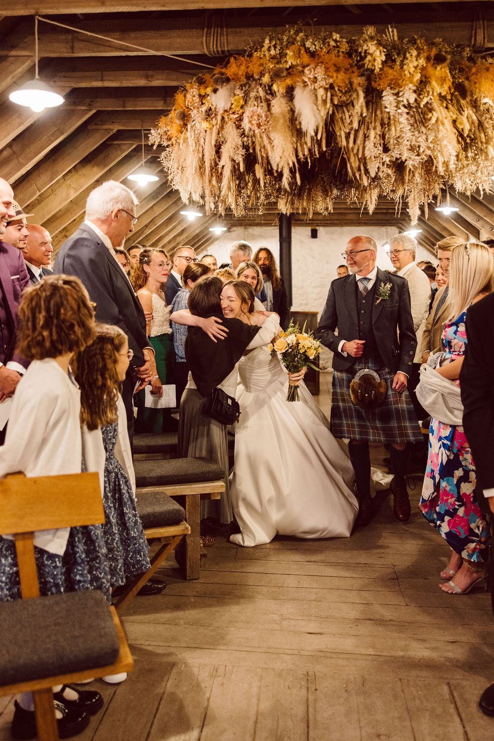 Wedding reception guests dance and celebrate in a rustic barn venue decorated with hanging dried flowers.