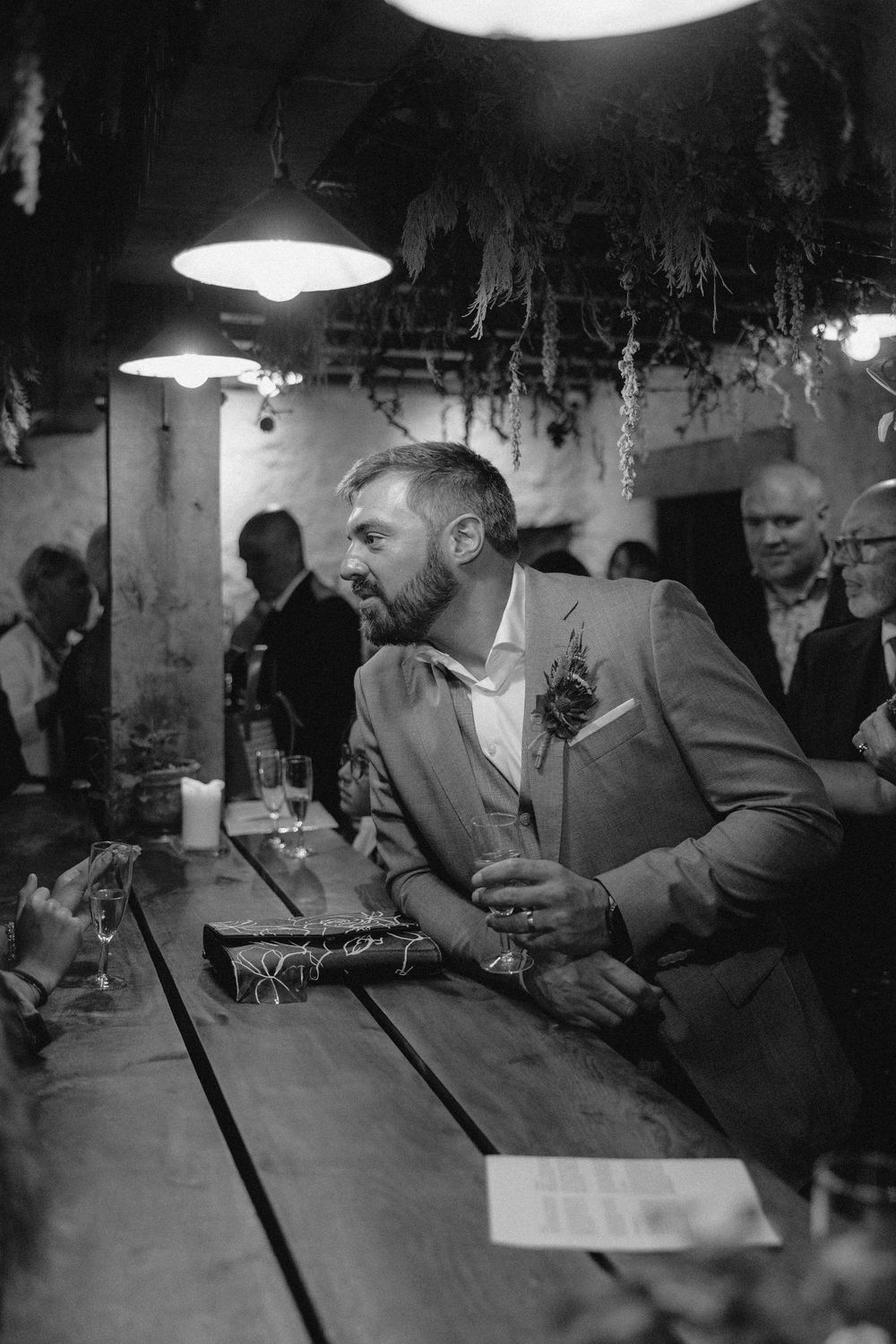 Black and white image of patrons sitting at a dimly lit bar counter with overhead pendant lighting.