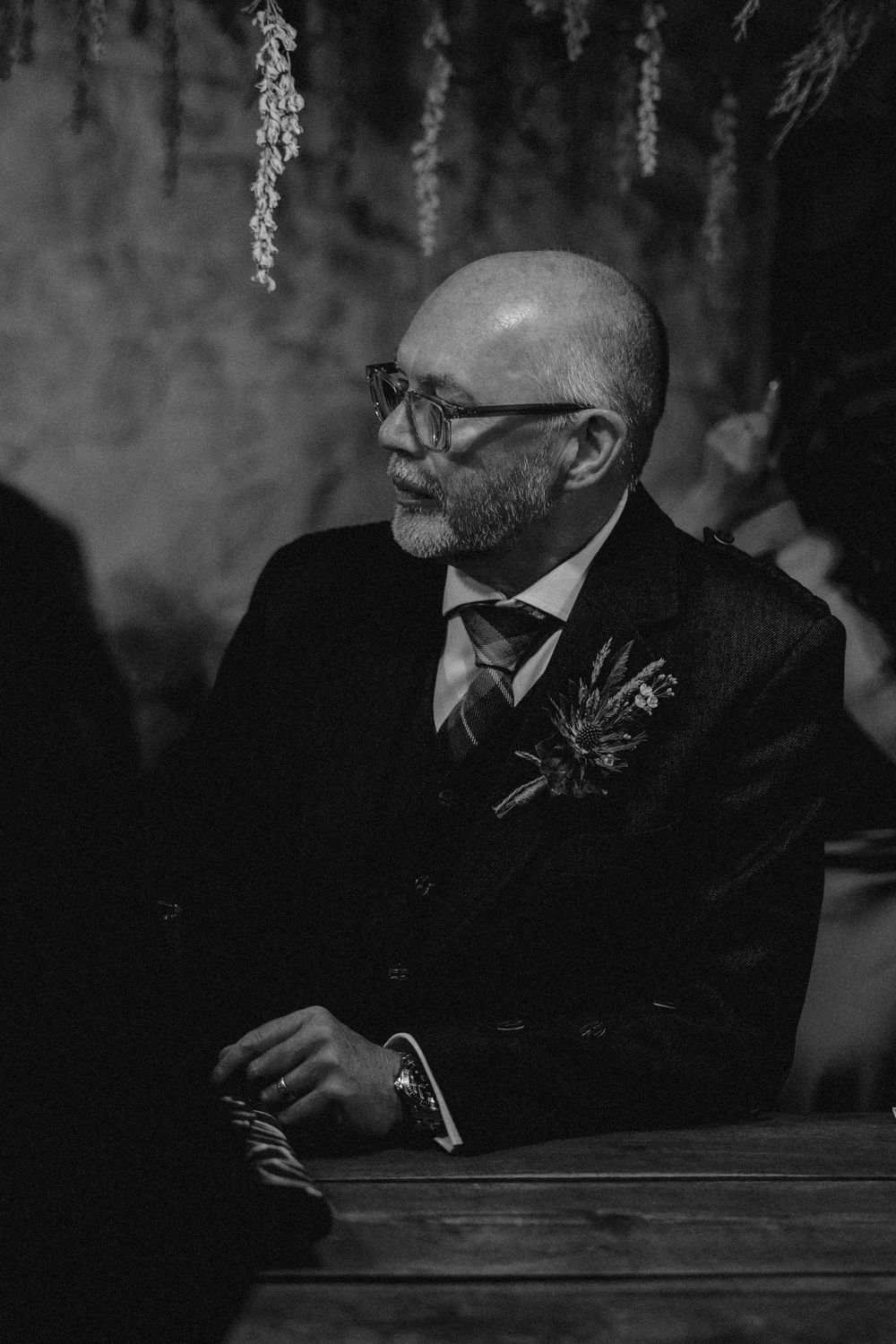 Black and white portrait of a bald man in glasses and formal suit with boutonniere sitting at wooden table.