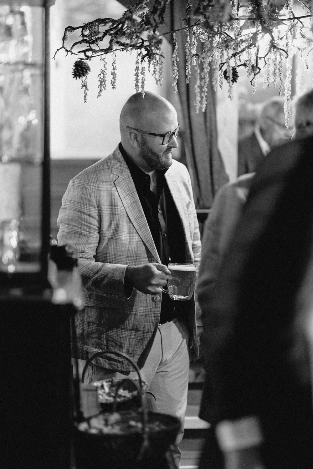 A man in a checkered blazer holds a drink while mingling at an elegant indoor event.