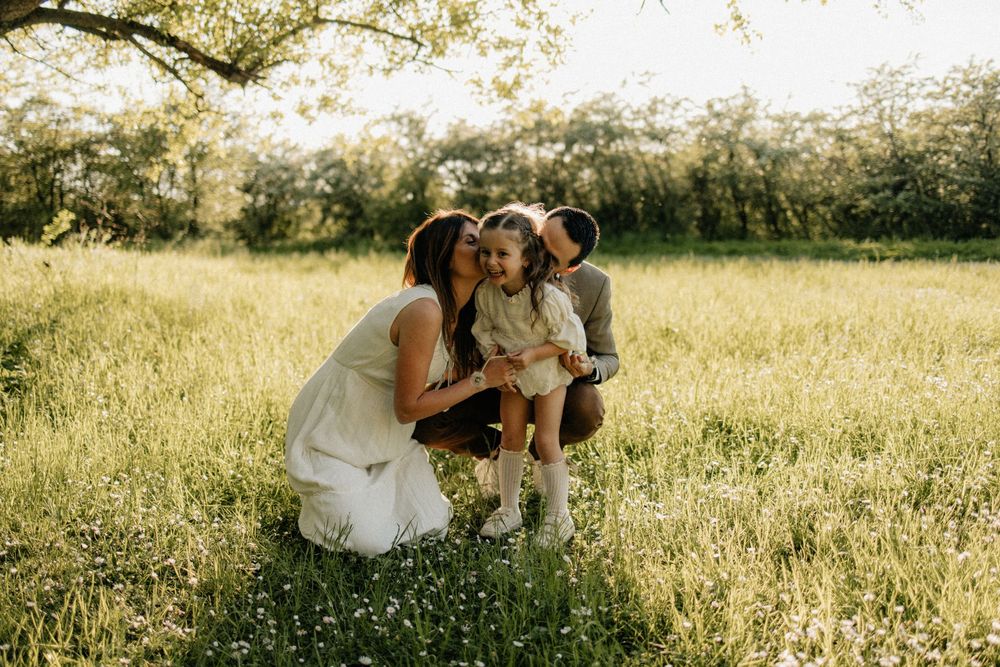 A family shares a tender moment together in a sun-drenched meadow with tall grass and wildflowers at golden hour.