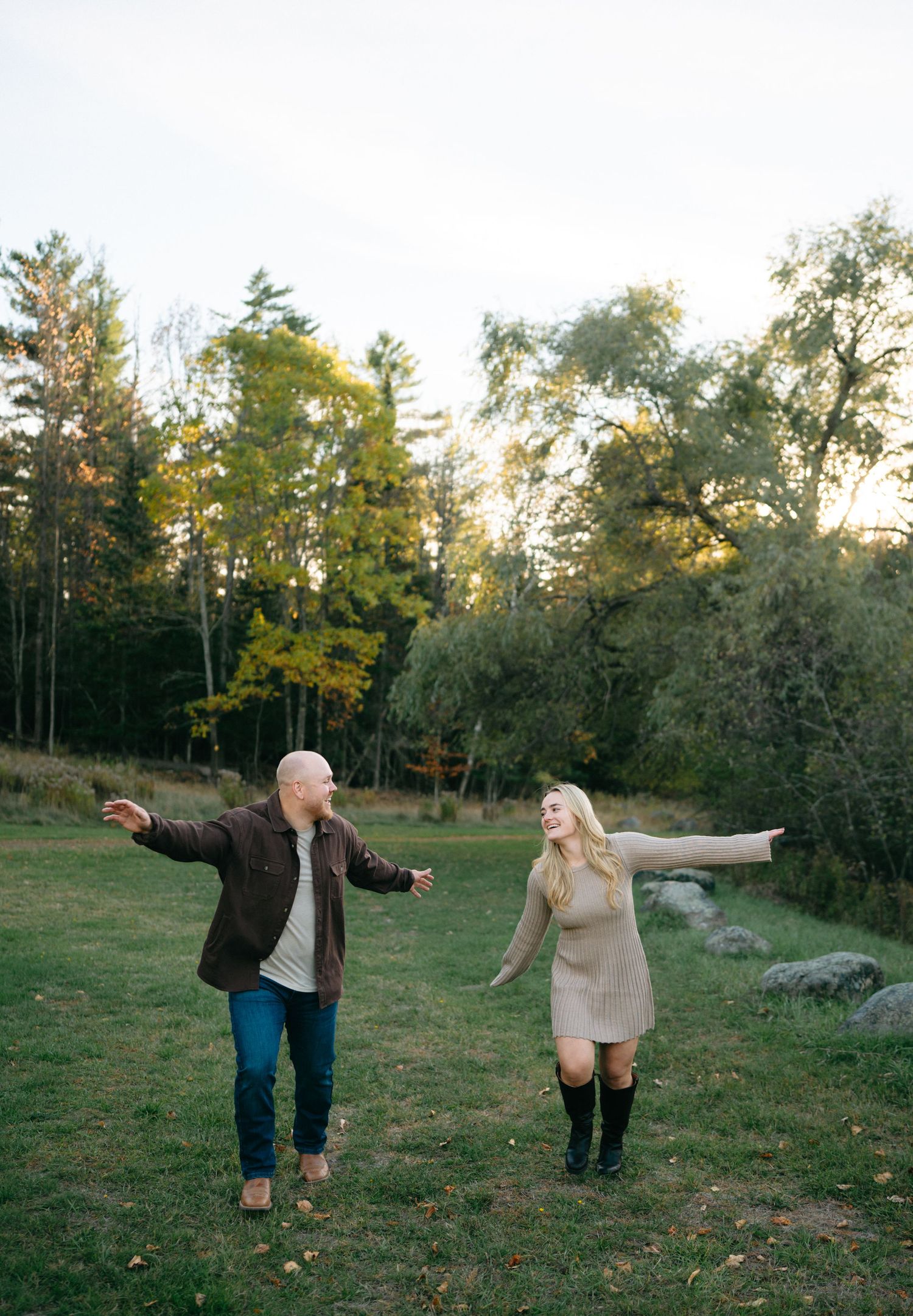 A couple dances playfully together in an autumn field surrounded by trees during golden hour.