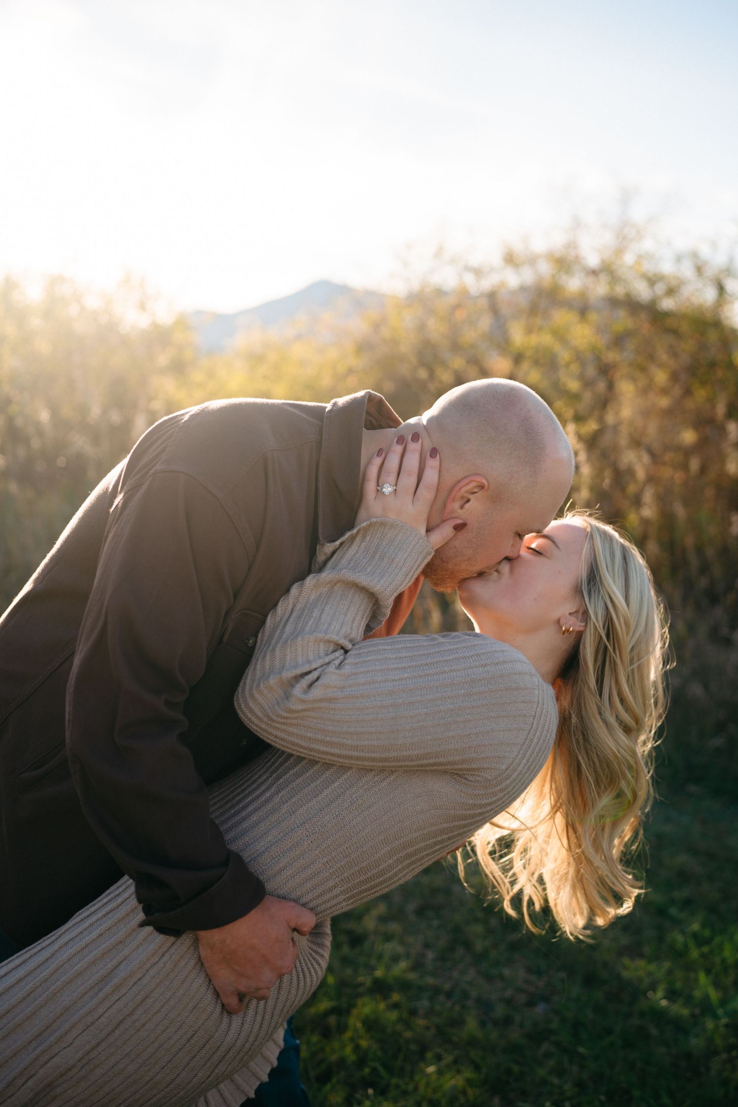 A romantic couple embraces and kisses in a natural outdoor setting with golden sunlight filtering through trees.
