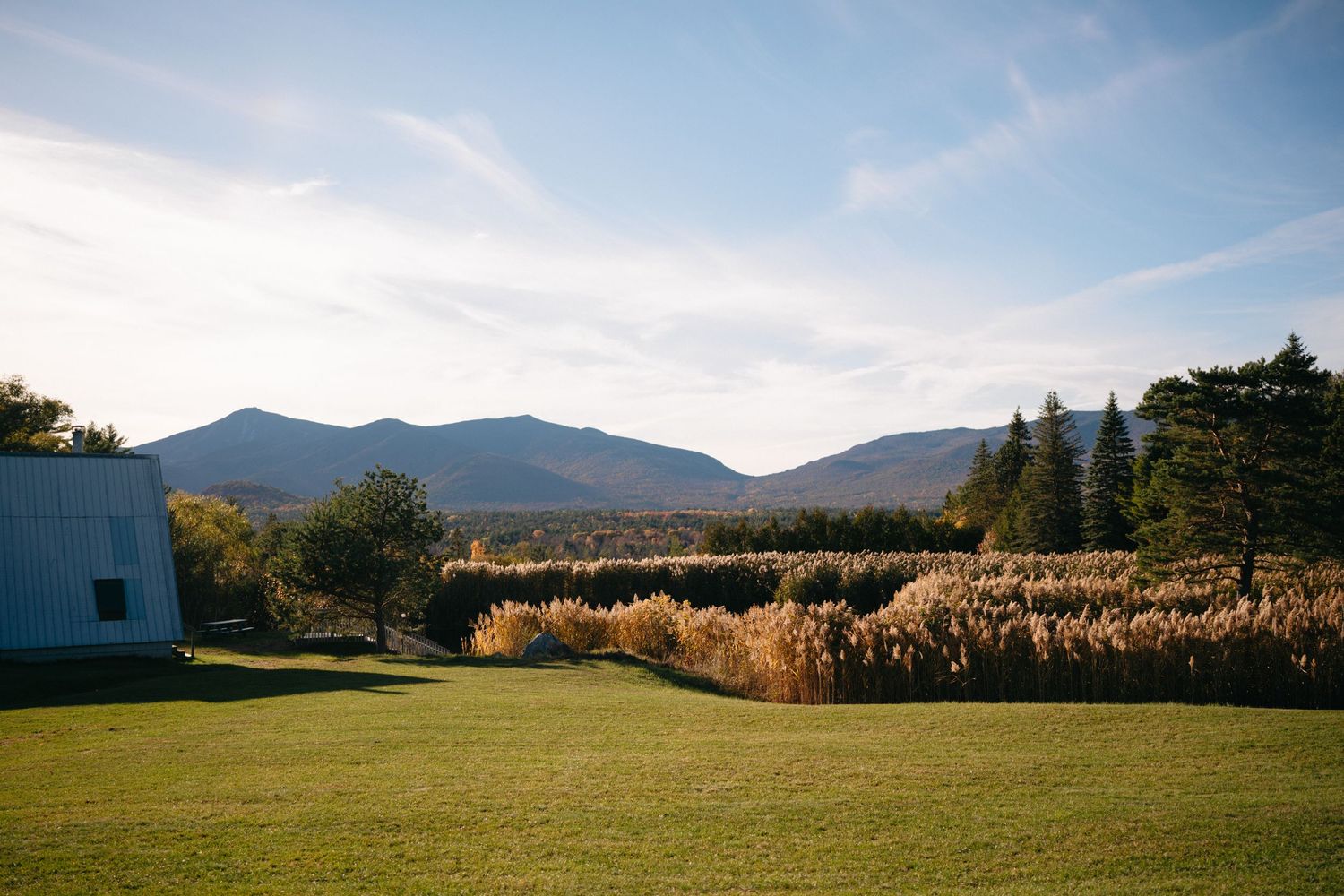 A scenic mountain landscape with a grassy lawn and golden field against blue sky and rolling hills in the background.