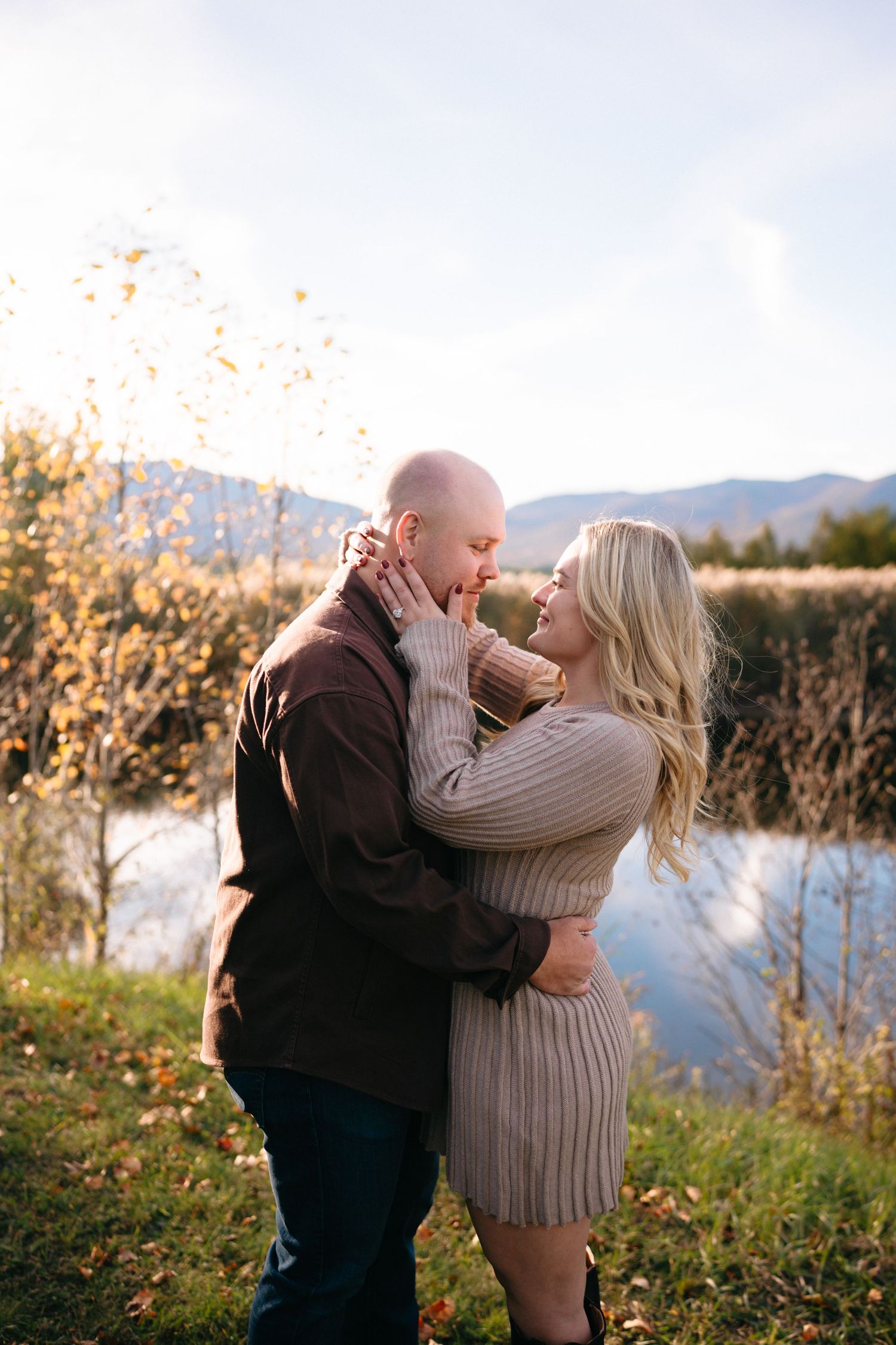 Couple embraces during a romantic sunset proposal moment by a lake with mountains in the background.