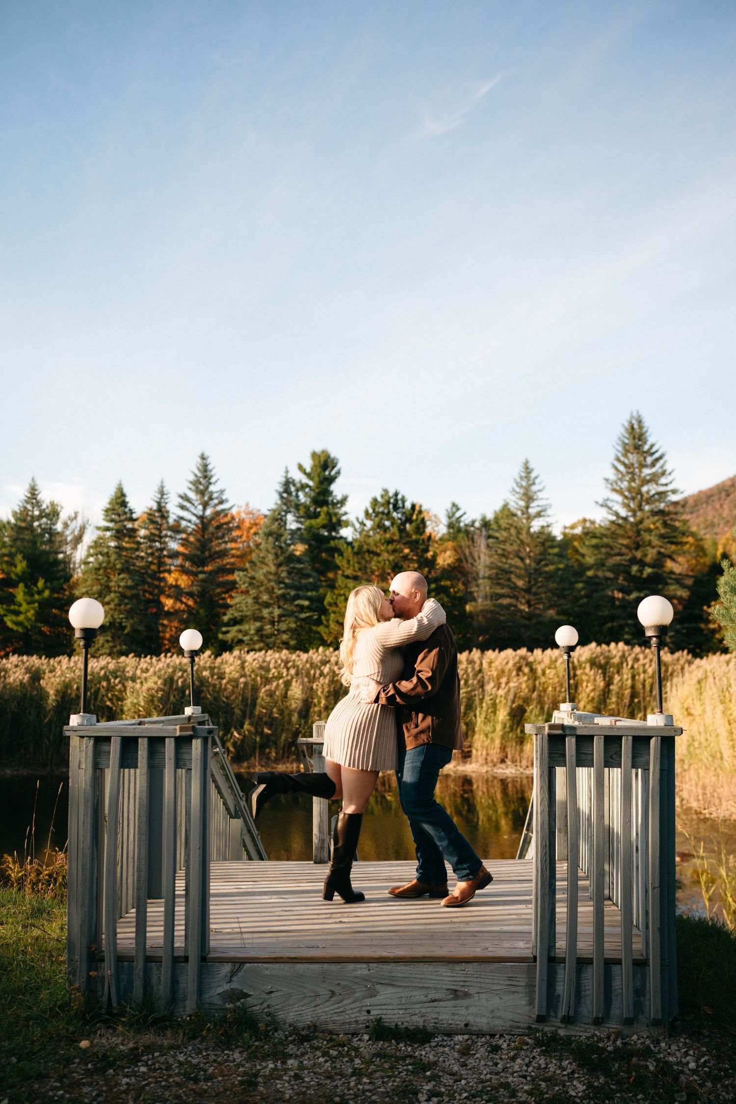 A couple shares a romantic moment on a wooden dock surrounded by evergreen trees and tall grass at sunset.