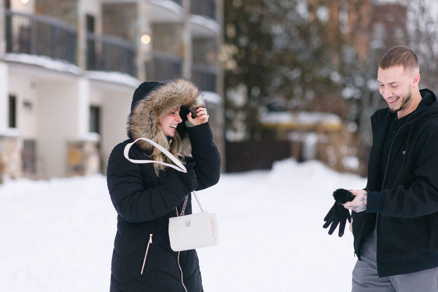 Two people in winter coats share a playful moment while standing in the snow outside a building.