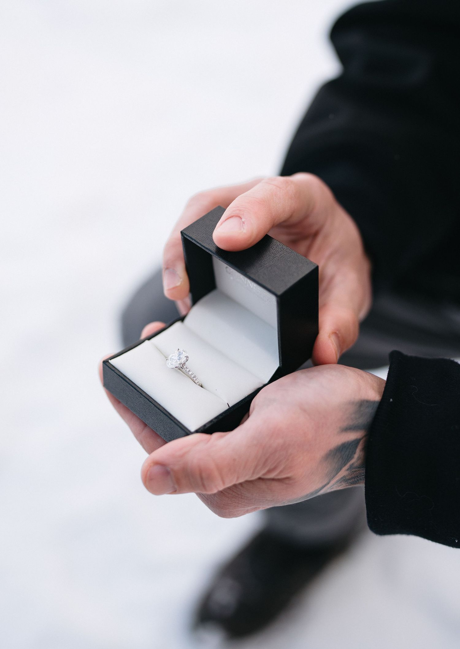 Hands holding a small black jewelry box with a diamond engagement ring inside, shown from multiple angles.