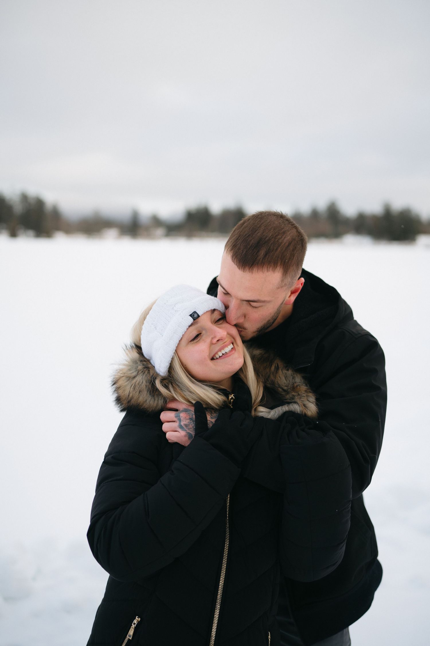 A couple in winter coats share a tender moment while standing in a snowy landscape.