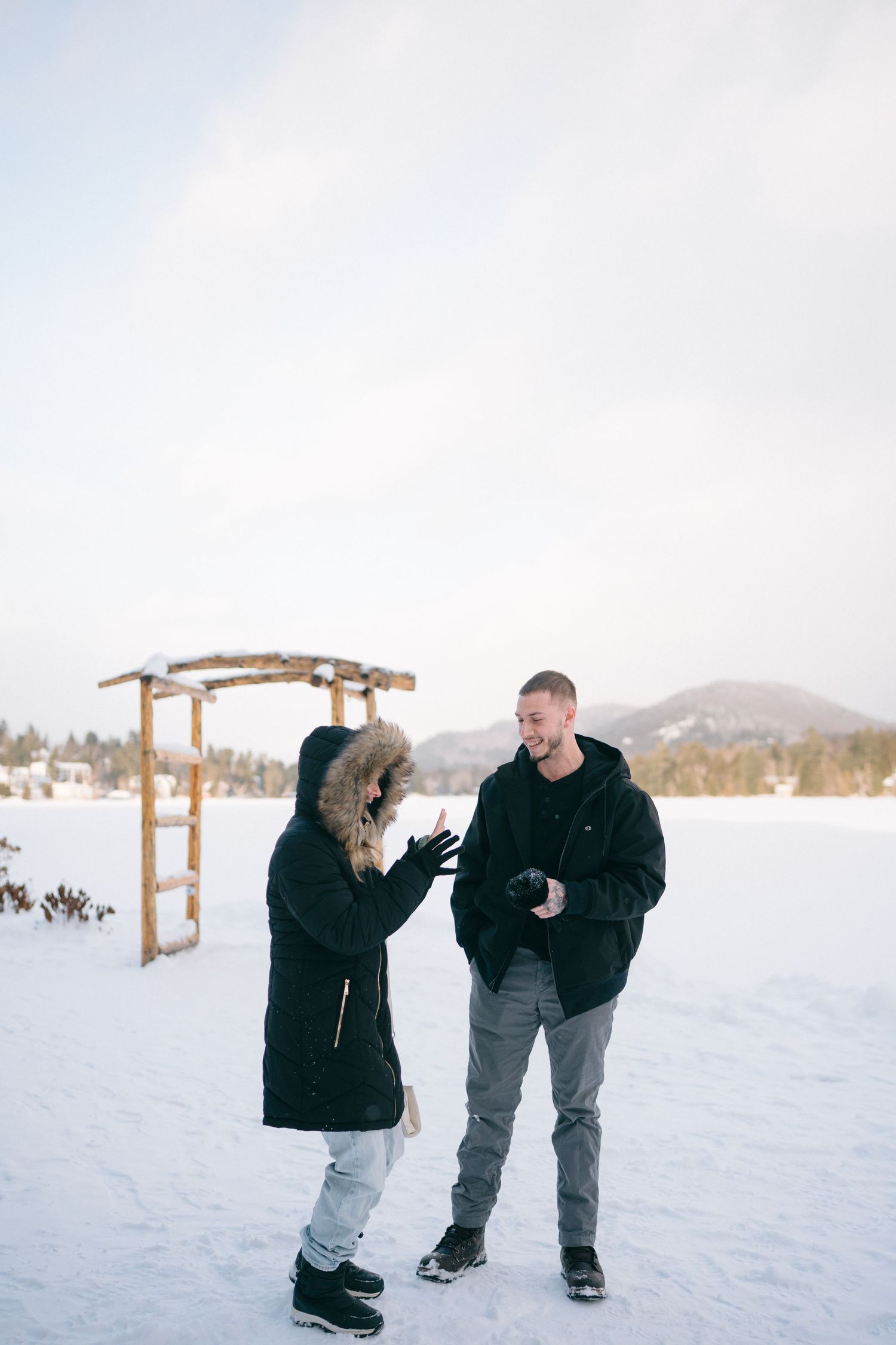 A couple in winter coats shares a playful moment in the snow with mountains visible in the background.