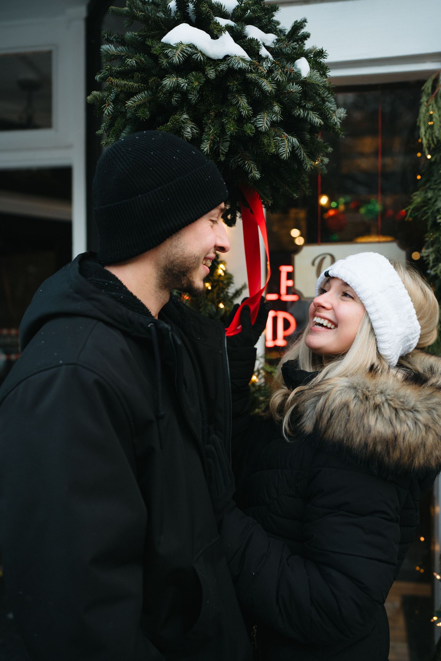 A couple shares tender moments under evergreen branches with holiday lights in the background.