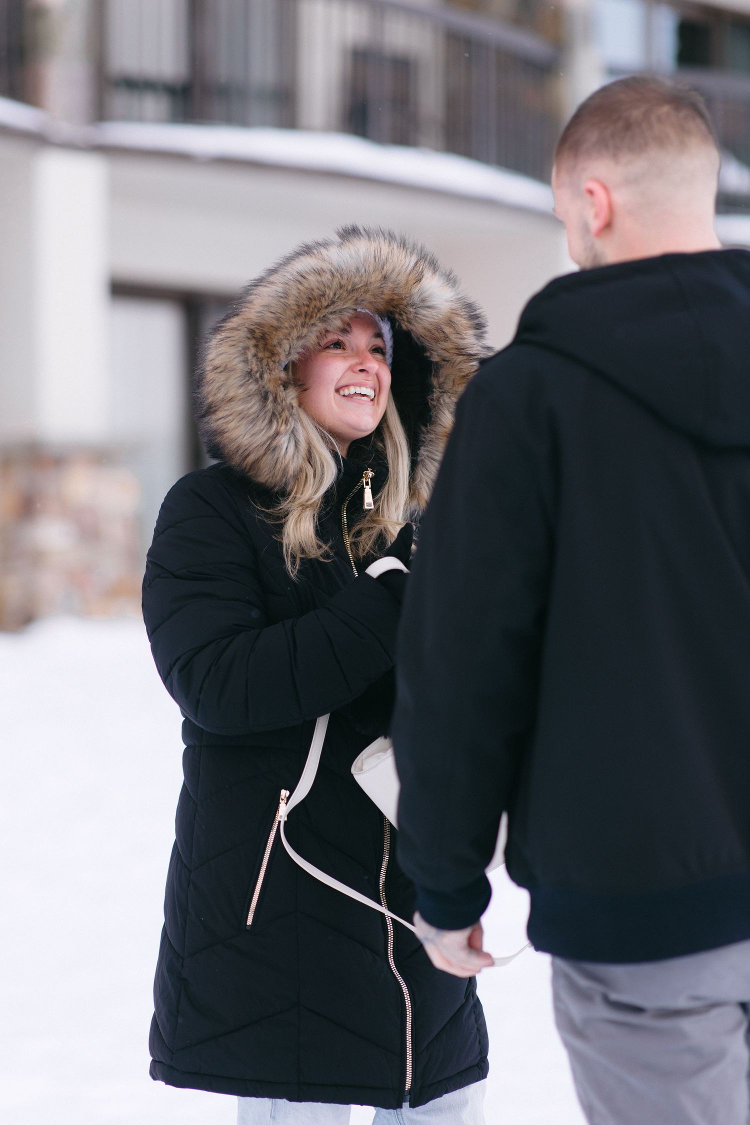 A couple shares a joyful moment outdoors in winter, with one person wearing a black fur-hooded parka coat.