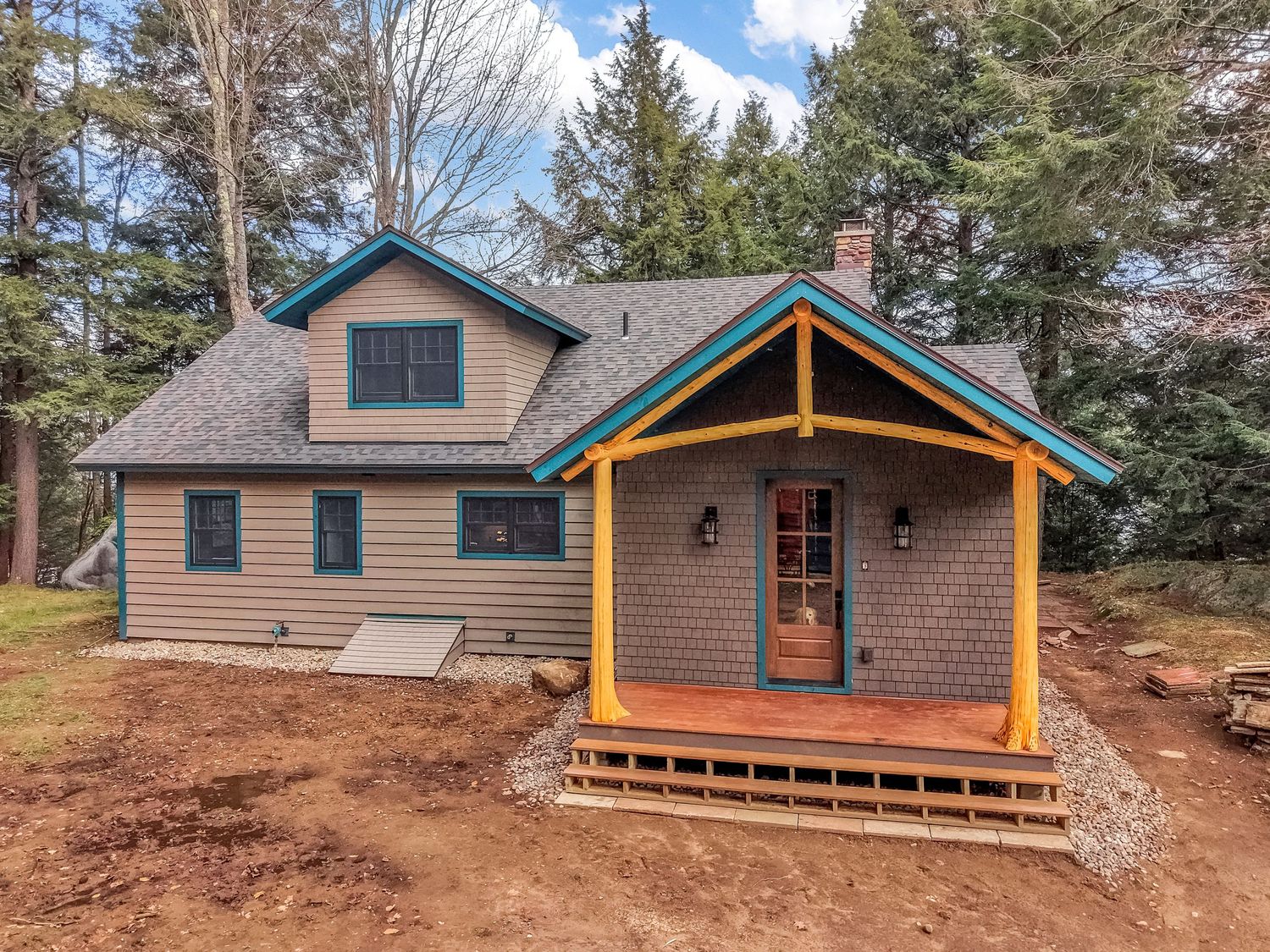 Modern mountain cabin exterior with teal trim and wooden deck nestled among tall pine trees in a forest setting.