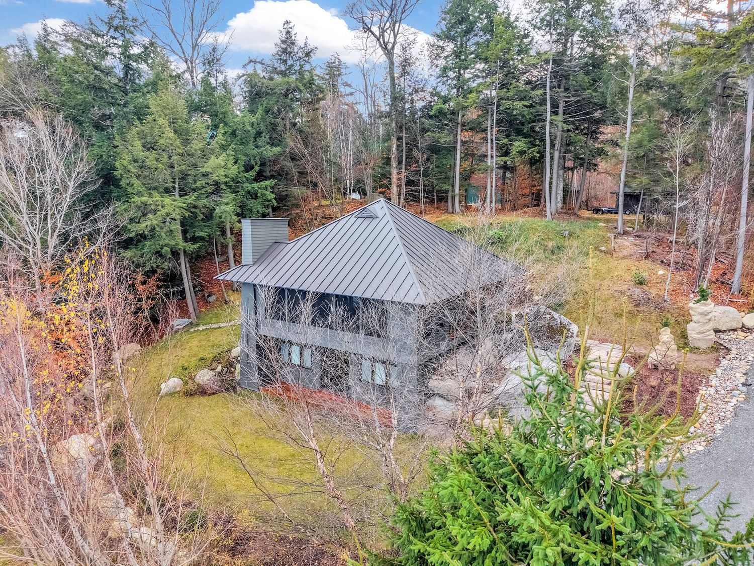 Aerial view of a modern gray house nestled among evergreen trees and autumn foliage in a wooded mountain setting.