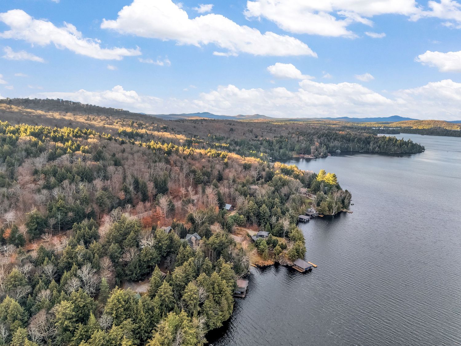 Aerial panoramic view of a scenic lake surrounded by autumn forest and mountains under blue skies with white clouds.