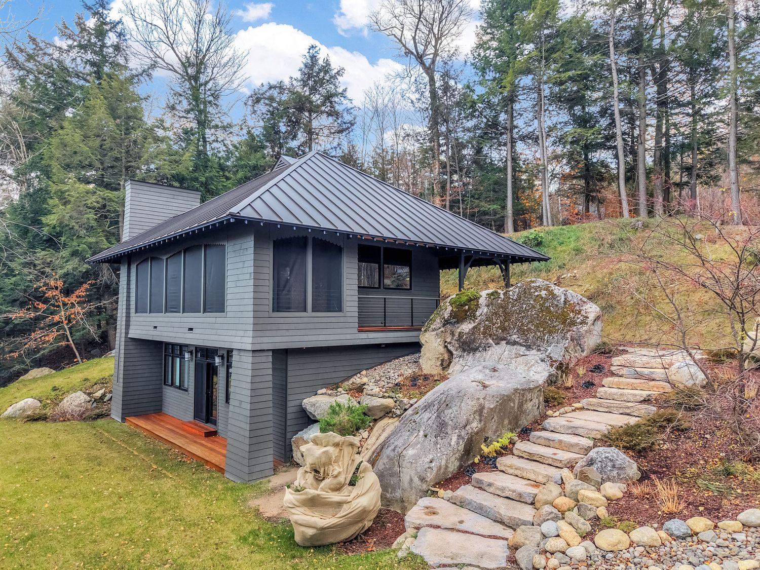 Modern gray house with metal roof perched on hillside featuring stone steps and natural landscaping in wooded setting.