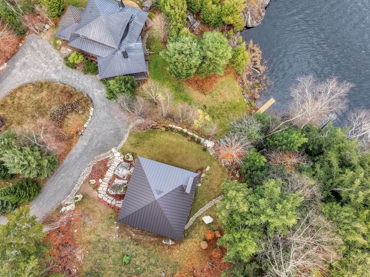 Drone view of waterfront vacation homes surrounded by evergreen trees and autumn foliage along a peaceful lake.