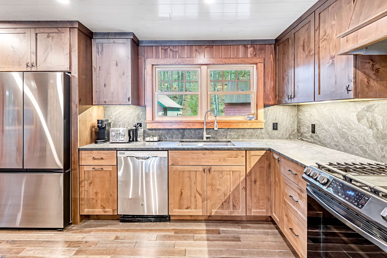 Modern kitchen with wood cabinets, stainless appliances, and large window overlooking nature.