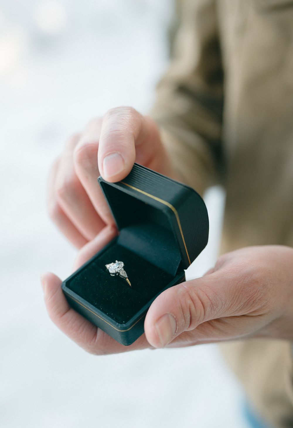 Hands holding open a dark blue velvet ring box with a sparkling diamond engagement ring inside.