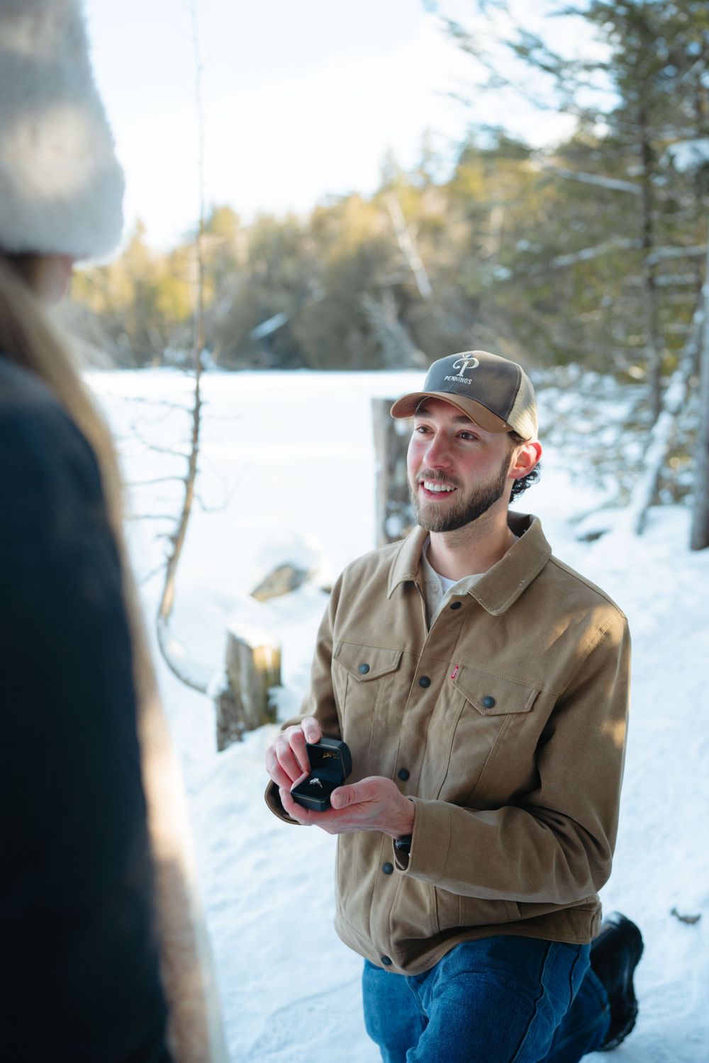 A photographer in a tan jacket and baseball cap kneels in the snow while taking pictures on a winter day.
