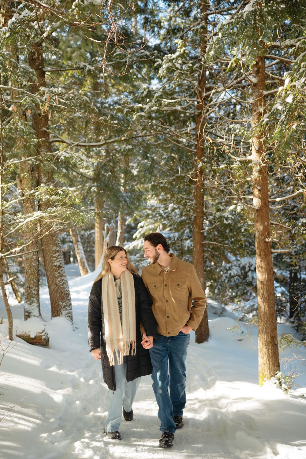 A couple walks hand in hand through a snowy forest path surrounded by tall pine trees on a sunny winter day.
