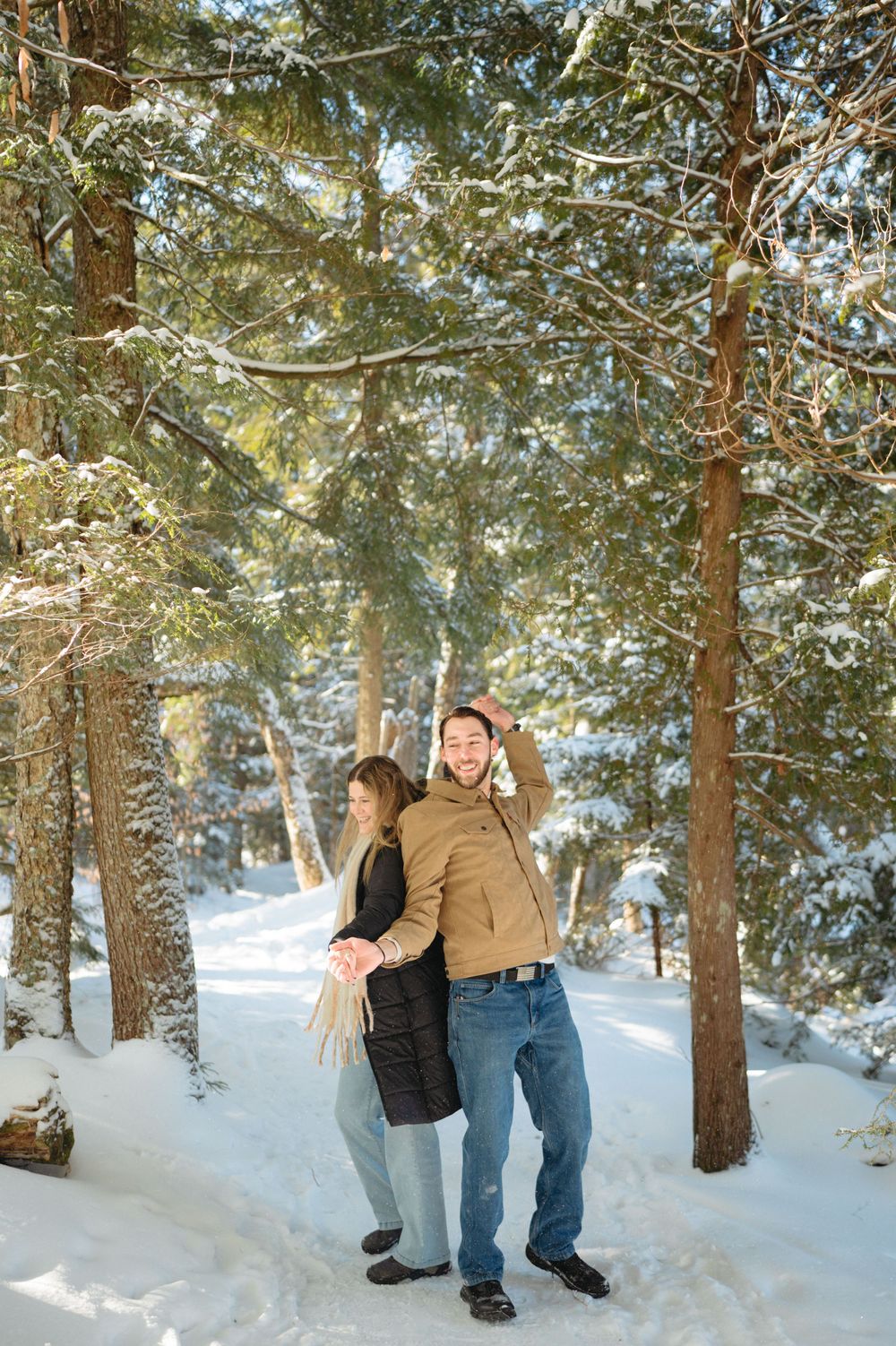 A couple enjoys a romantic winter stroll through a snow-covered forest path surrounded by tall evergreen trees.