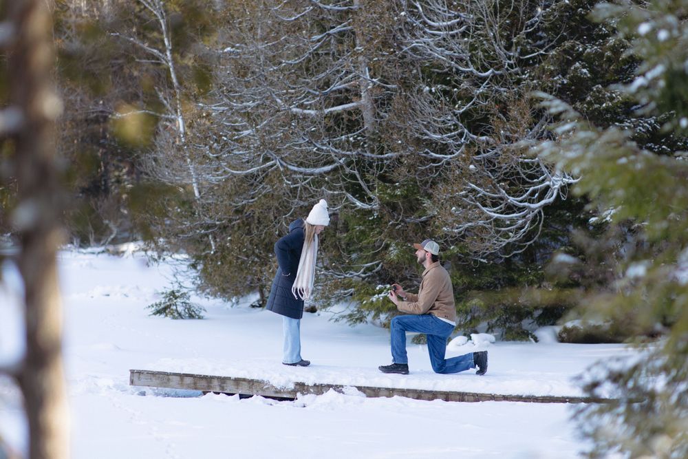 A couple in winter coats stands on a snowy wooden deck surrounded by evergreen trees during a marriage proposal.