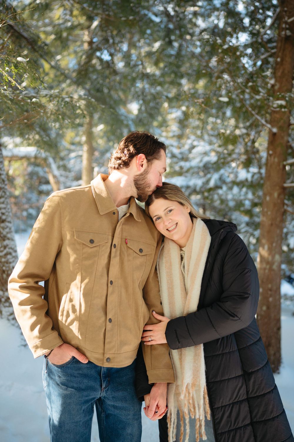 Couple shares intimate moments in a snowy winter forest setting wearing tan and black coats.