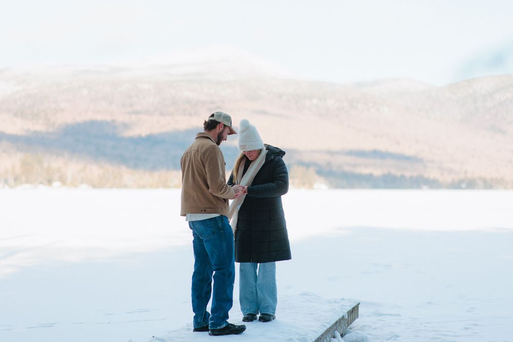 A couple stands together on a snowy mountain overlook during winter, with scenic views of distant peaks and valleys.