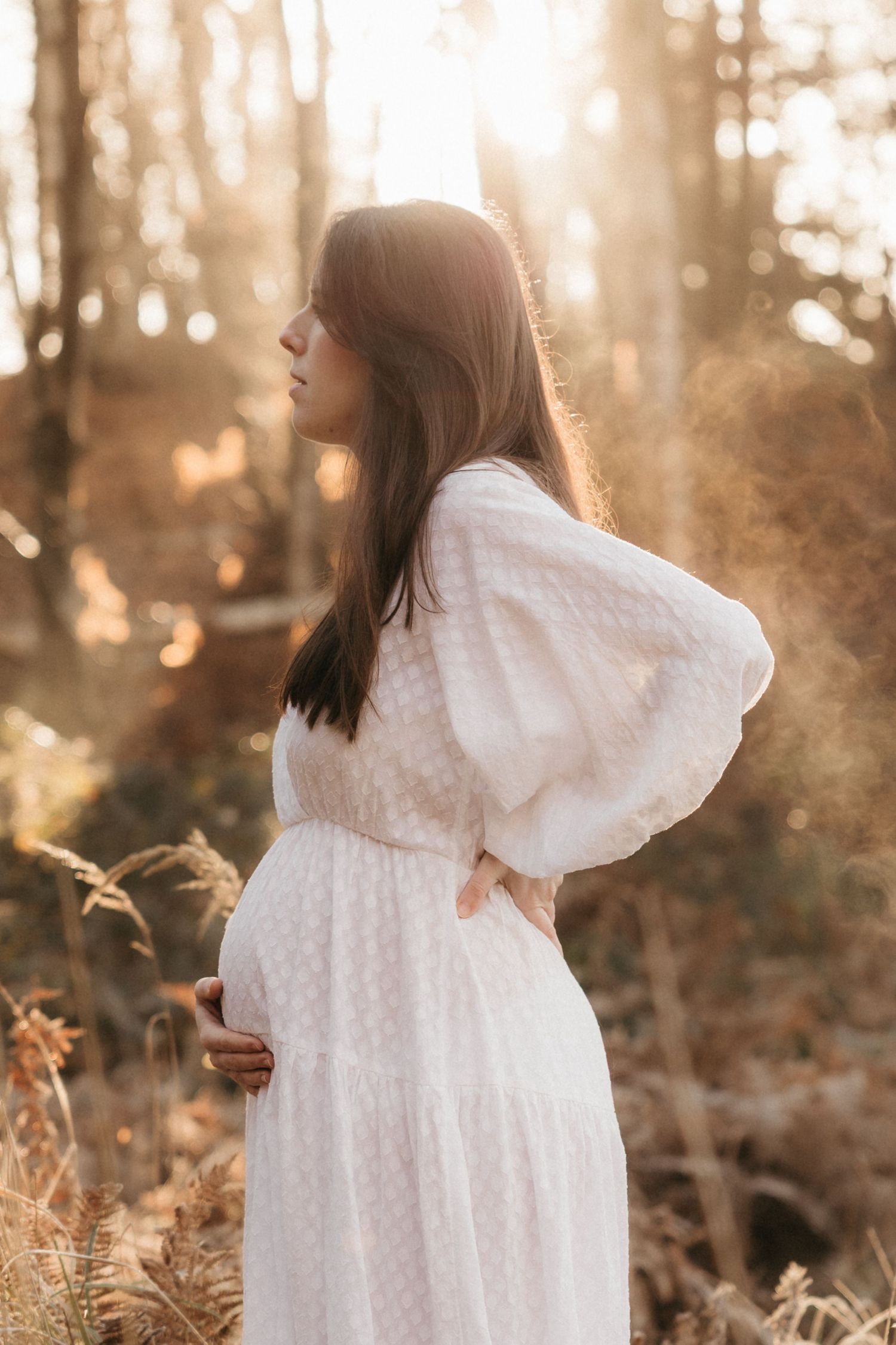A figure in a flowing white dress stands in a sunlit forest with golden light filtering through the trees.