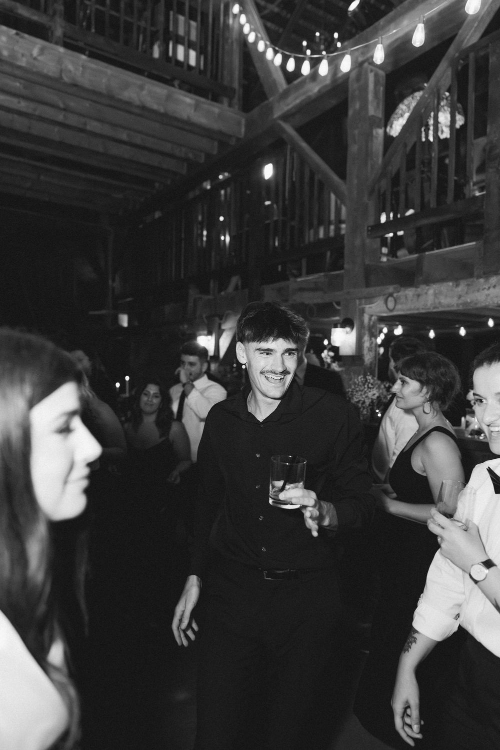 Wedding guests dancing and celebrating in a rustic barn venue with string lights overhead.