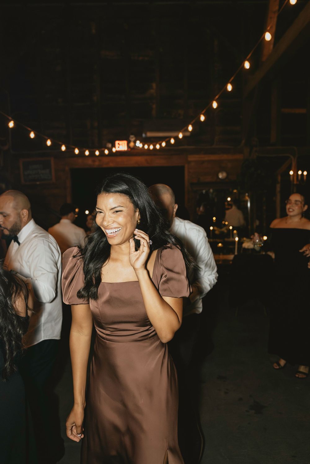 Wedding guest in brown satin dress smiles joyfully under string lights at evening reception celebration.