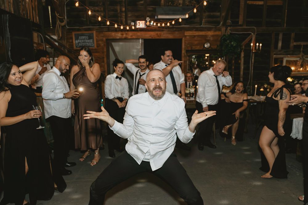 Wedding guest shows off dance moves in white shirt on rustic barn dance floor with string lights.
