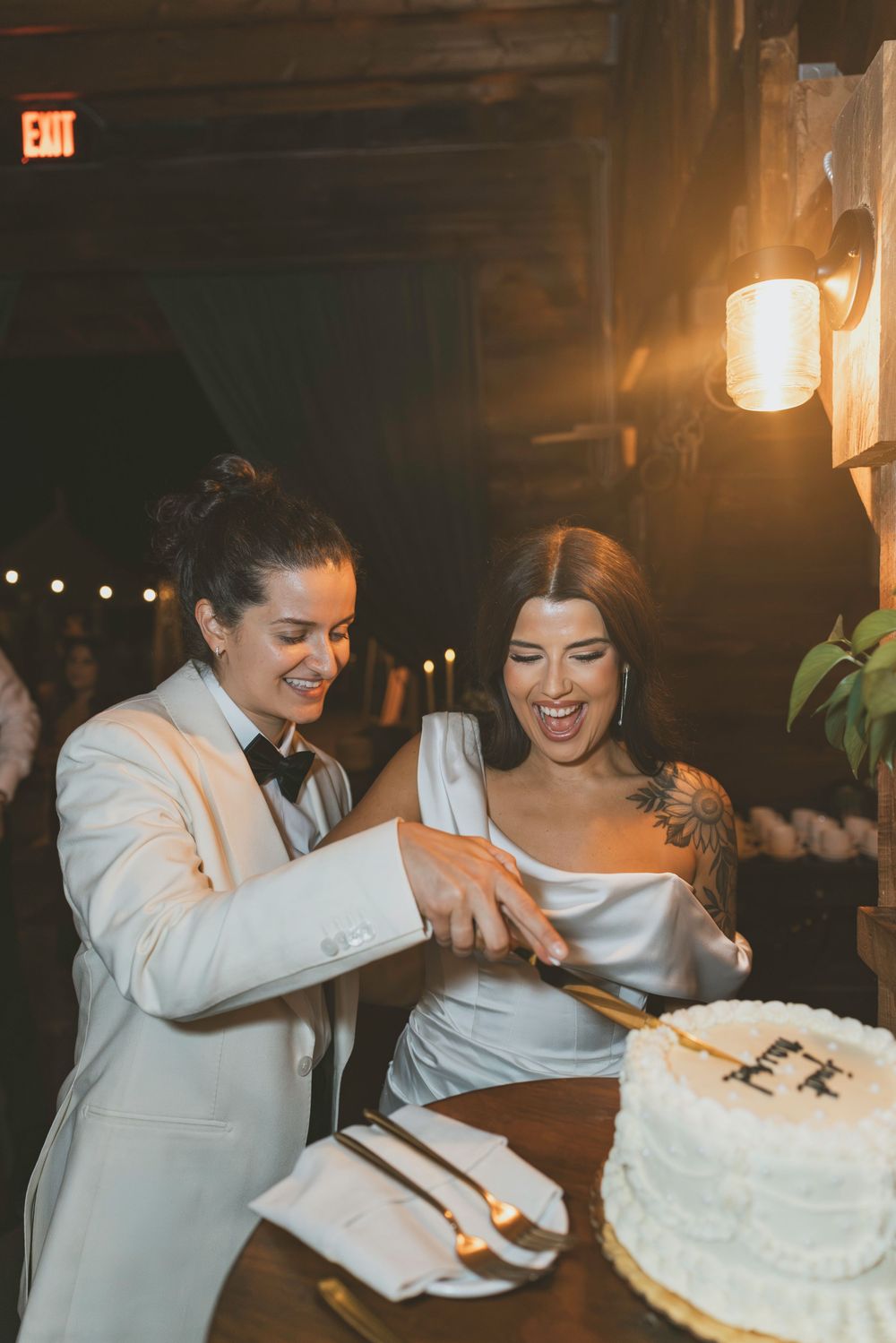 Couple cuts their wedding cake together at rustic barn reception under warm lighting.