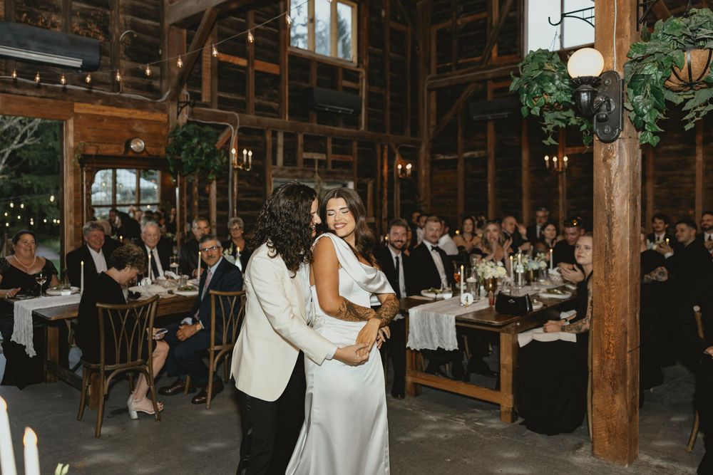 Wedding guests dance and celebrate on a rustic wooden dance floor under string lights in a barn venue.