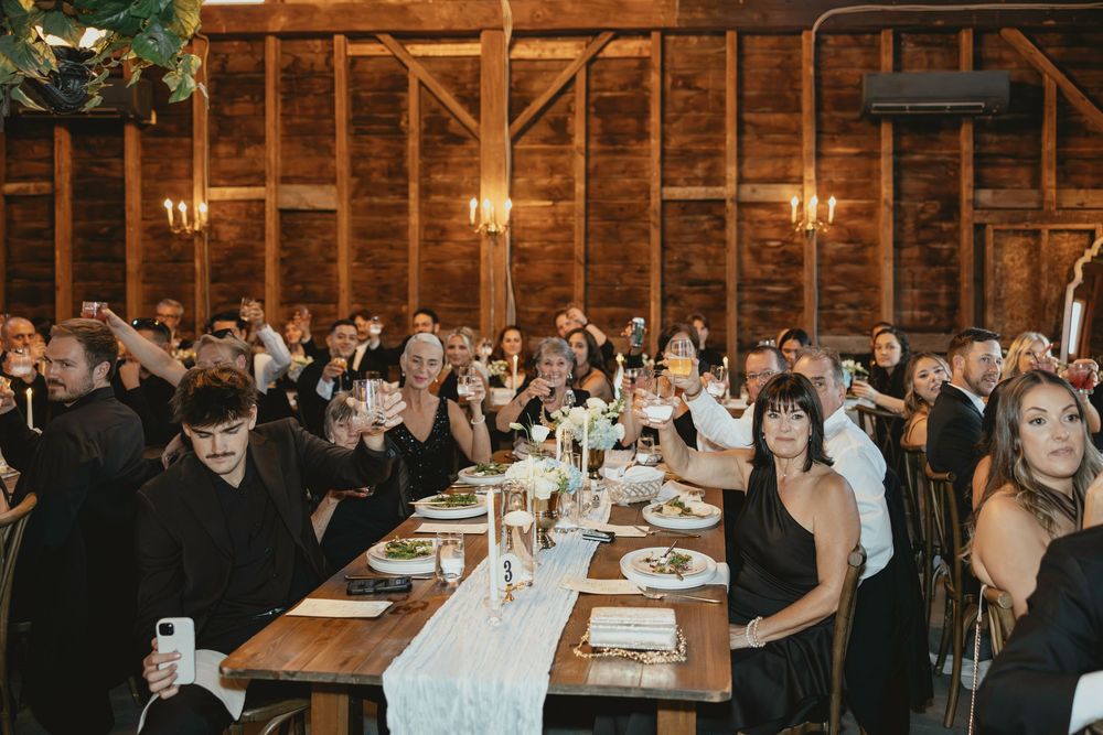 Wedding reception guests seated at long wooden tables in a rustic barn venue with warm lighting and elegant decor.