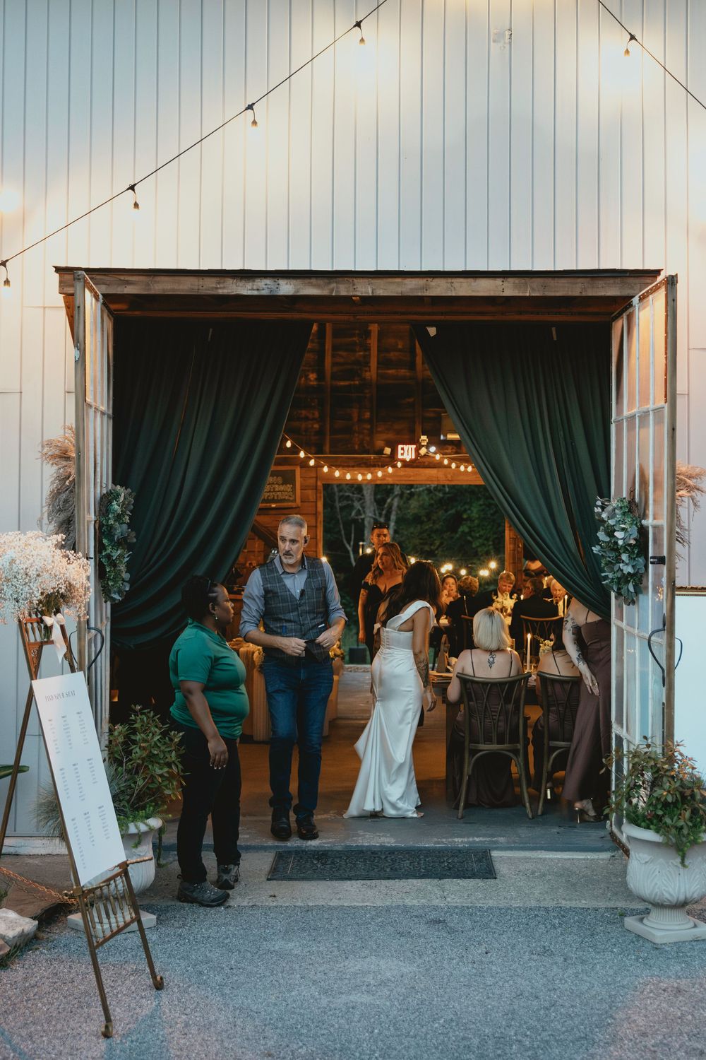 Wedding guests gather at a rustic barn venue entrance decorated with string lights and elegant curtains.