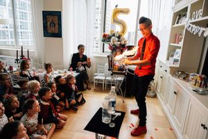 A magician performs tricks for a group of seated children at a birthday party in a bright living room.