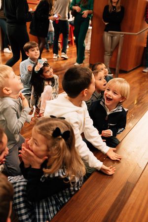 Students sitting at wooden benches laugh and react with excitement during a school assembly or performance.