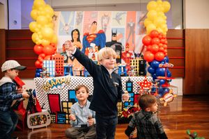 Children celebrating at a superhero themed birthday party with yellow and red balloon decorations and colorful backdrop.