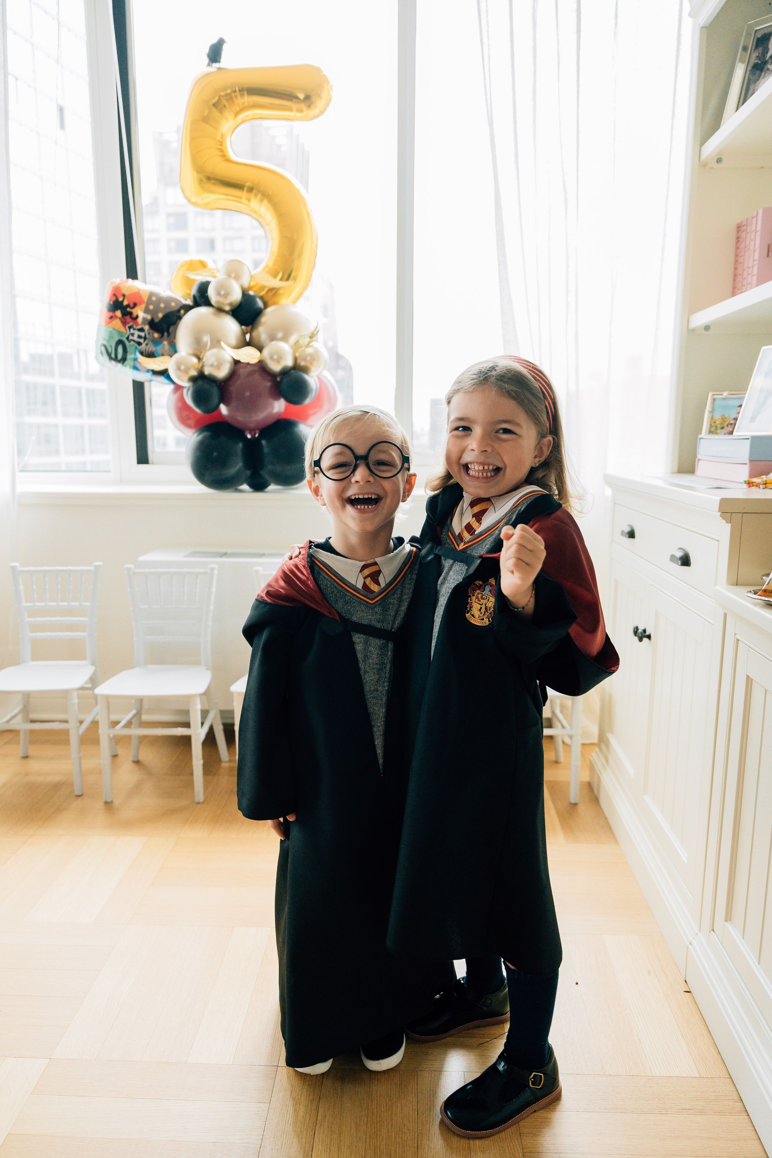 Two children in matching black outfits pose playfully with gold number balloons in a bright white room.
