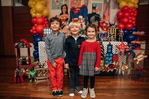 Three children pose in front of superhero party decorations with balloon columns and wrapped presents.