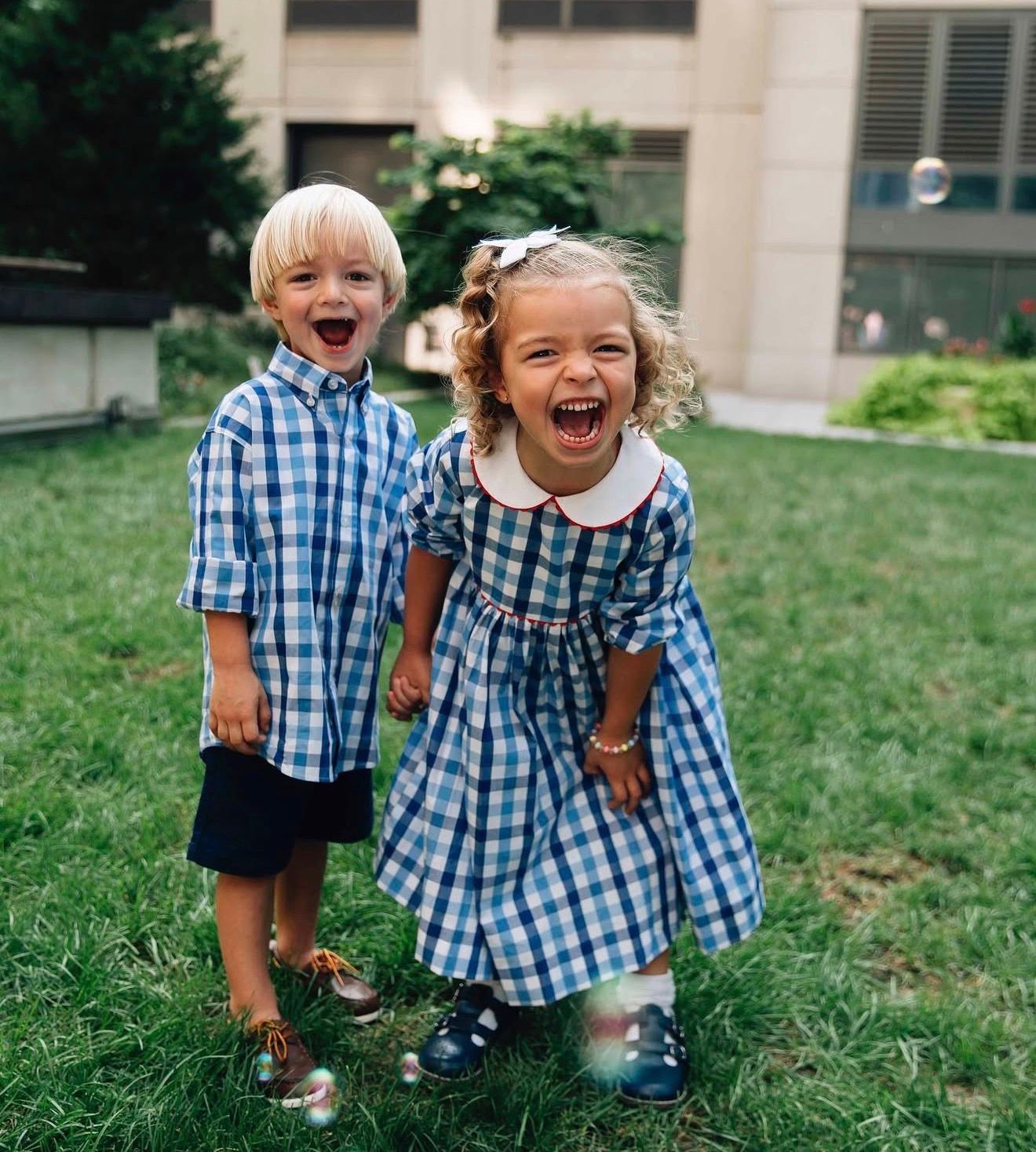 Two young children in matching blue checkered outfits laugh joyfully while playing on grass outside a building.