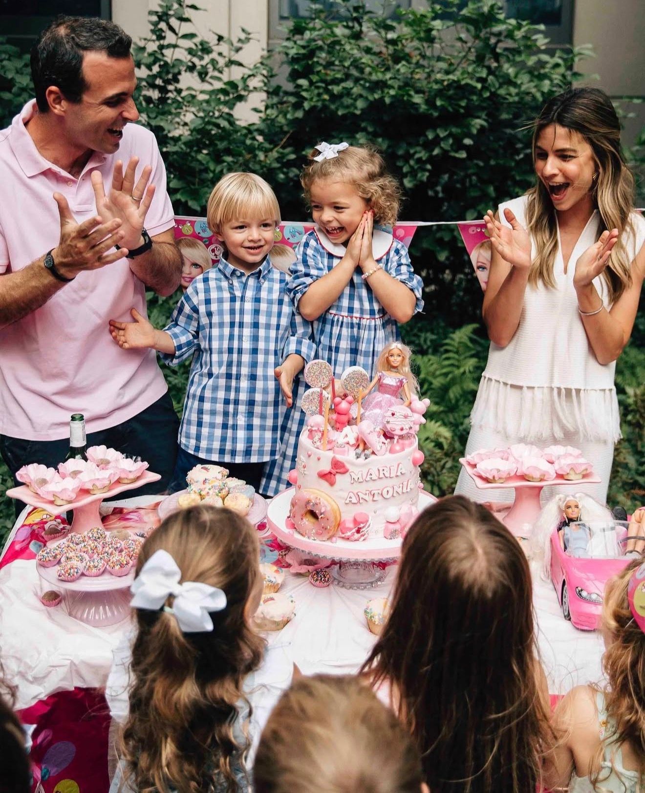 A group of people enjoying a birthday celebration with pink cake and laughter in an outdoor garden setting.