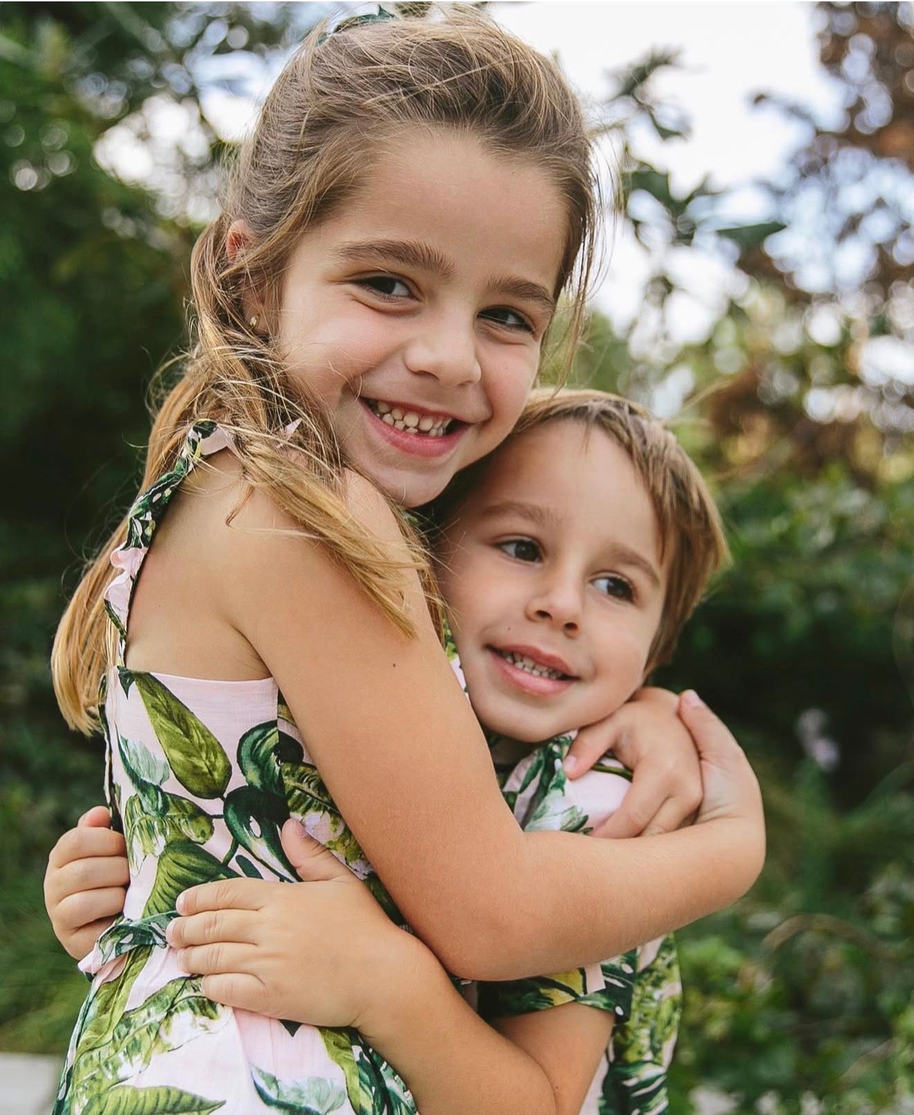 Two young children share a warm embrace outdoors while wearing floral print clothing.
