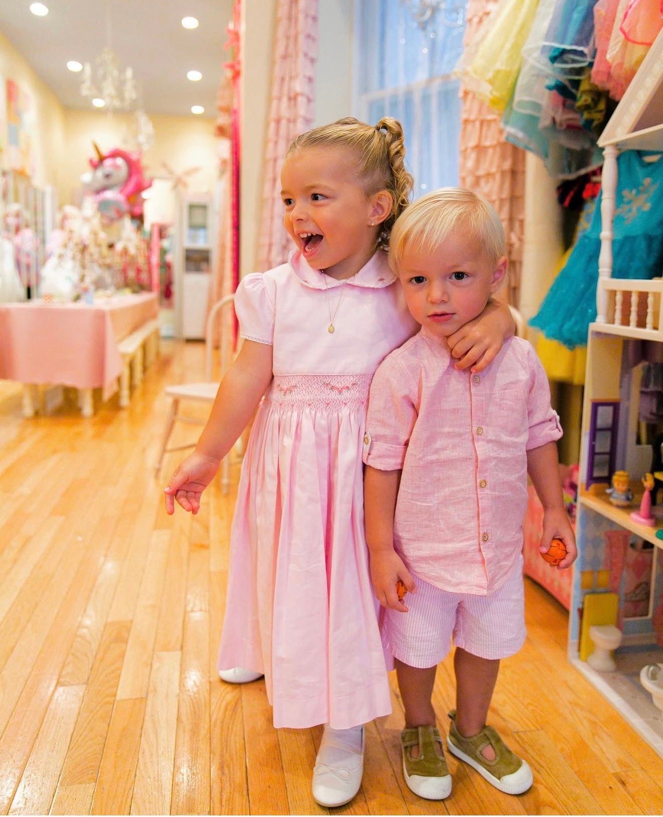 Two small children in matching pink and white outfits hold hands while walking through a brightly lit retail store.