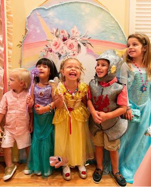 Children in colorful princess dresses and party outfits laugh together while lined up against a wall at a celebration.