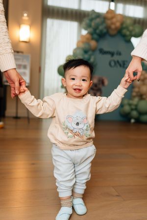 A baby in a koala sweater and white pants holds hands with adults while learning to walk on hardwood floors.